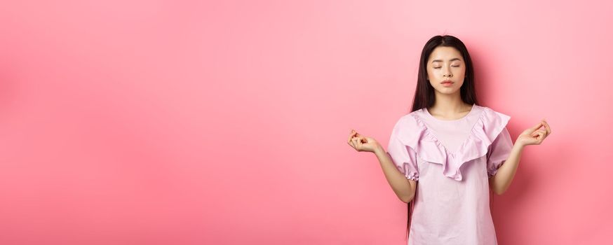 Stay calm. Peaceful asian woman meditating, hold hans in zen gseture and close eyes, practice yoga, standing patient on pink background.