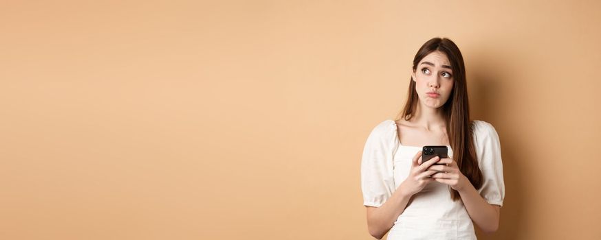 Confused young woman thinking after reading news on mobile phone, looking at upper left corner hesitant, standing on beige background.