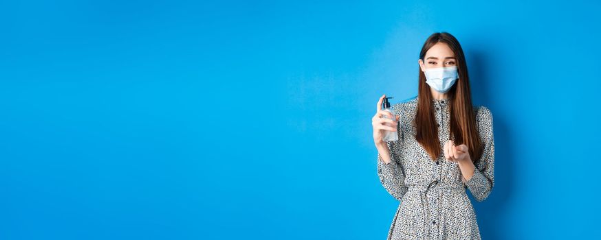 Covid-19, social distancing and healthcare concept. Beautiful girl in medical mask using hand saniziter, holding antiseptic bottle, standing on blue background.