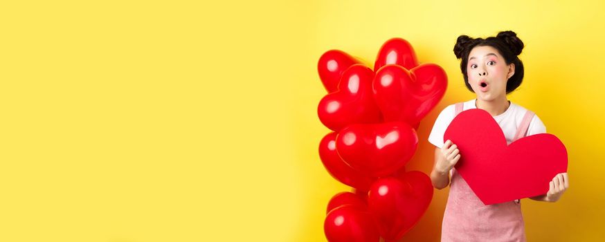 Happy Valentines day. Silly asian girl showing big red heart postcard and looking amazed, standing near red romantic balloons, yellow background.