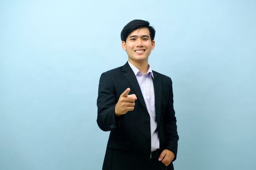 Portriat of asian young smart happy businessman dressed in suit standing straight, smiling, and pointing finger at camera with isolated light blue background. Business concept.