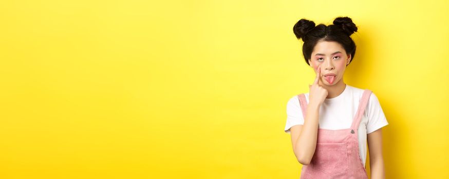 Rude asian girl stretch eyelid and showing tongue, mocking someone, standing on yellow background.