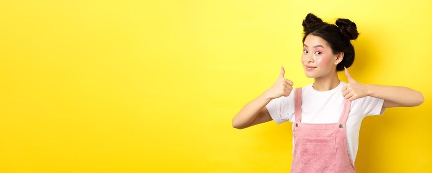 Beautiful asian girl with bright pink makeup, showing thumb up in approval and smiling satisfied, recommending promo, yellow background.