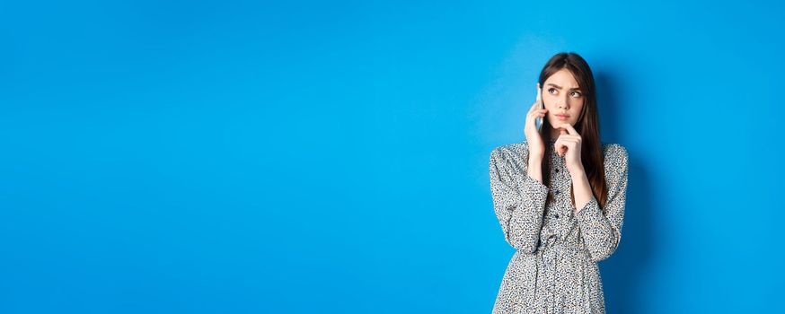 Pensive woman talking on phone and thinking, looking aside thoughtful, standing against blue background.