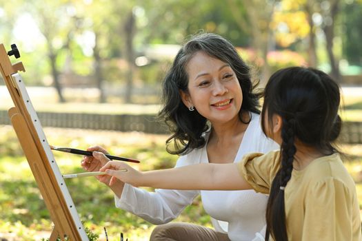 Positive mature grandmother and little granddaughter painting picture, enjoying leisure weekend hobby activity together outdoor.