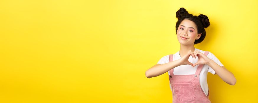 Valentines day. Cute asian girl sending her love, showing heart gesture and smiling at camera romantic, standing on yellow background.