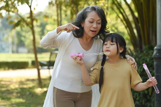Joyful little girl blowing soap bubbles with grandmother, spending leisure weekend time together at outdoor.