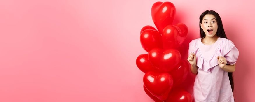 Excited and surprised teenage korean girl open mouth amazed, receive surprise gift on valentines day, looking wondered, standing near heart balloons, pink background.