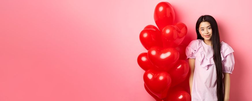 Cute and tender girl looking with love, celebrating valentines and white day with lover, standing near romantic heart balloons on pink background. Relationship concept.