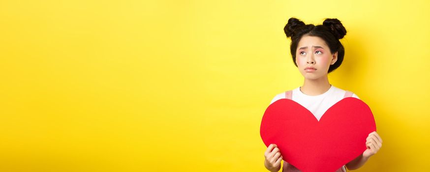Sad and lonely asian girl standing in make-up with big red heart cutout, left alone on Valentines day, looking left disappointed or upset, standing on yellow background.