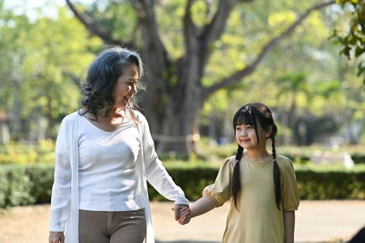 Smiling grandmother and her little grandchild walking together surrounded by trees during sunny day.