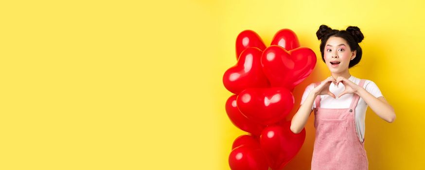 Happy asian girl showing heart sign near romantic balloons, waiting for true love on Valentines day, looking hopeful at camera, yellow background.