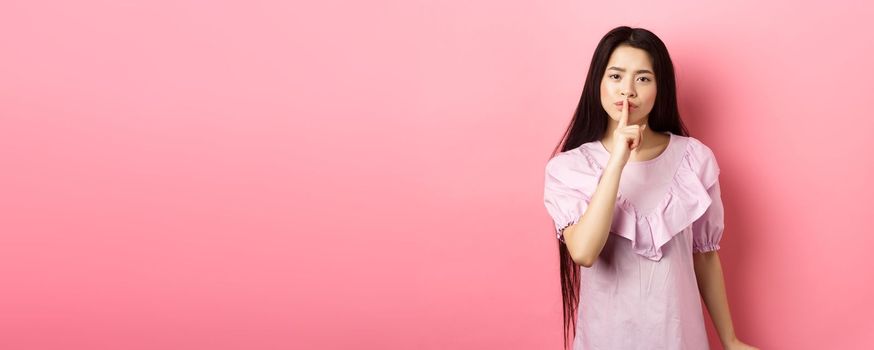 Cute asian girl tell to be quiet, scolding loud person, showing shush sign with finger pressed to lips, standing in dress on pink background.