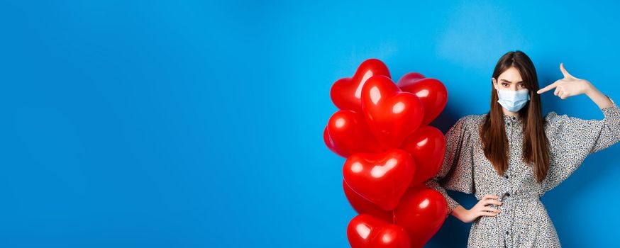 Covid-19, quarantine and health conept. Sassy young woman in medical mask, pointing finger at red hearts balloons and frowning, disappointed in Valentines day, blue background.