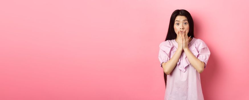 Shocked asian girl with long dark hair, gasping and covering mouth with hands, look at bad news, terrible accident, standing on pink background.