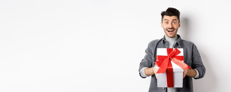 Surprised young man smiling excited, holding big gift box on valentines day holiday, receive surprise present, standing amazed on white background.