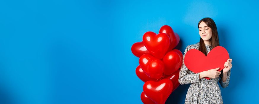 Valentines day. Romantic smiling woman hugging big red heart cutout and dreaming of love, standing in dress on blue background.