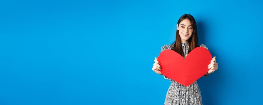 Valentines day and relationship concept. Beautiful romantic girl say I love you, extending hand with big red heart, standing on blue background.