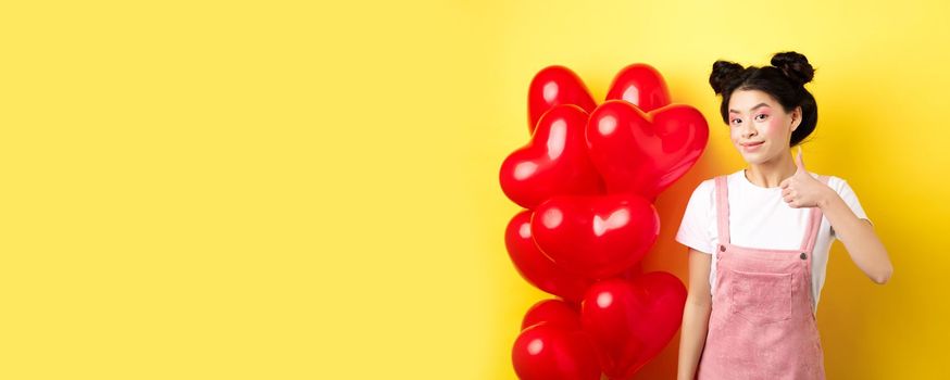 Fashionble asian woman in romantic outfit with make-up, showing thumb up and smiling, praising Valentines day offer, standing near red heart balloons, yellow background.