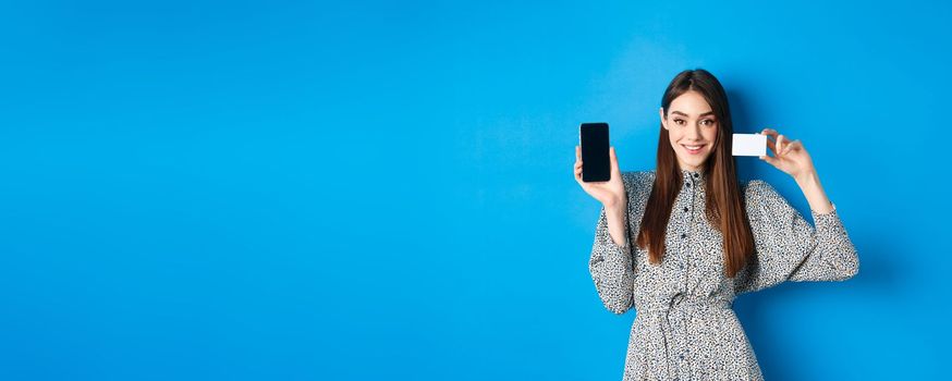 Online shopping. Smiling beautiful girl in dress showing plastic credit card and empty smartphone screen, blue background.