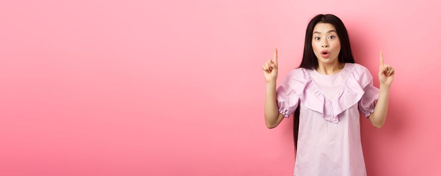 Excited asian girl showing awesome promo offer, pointing fingers up and gasping amazed, standing on pink background.