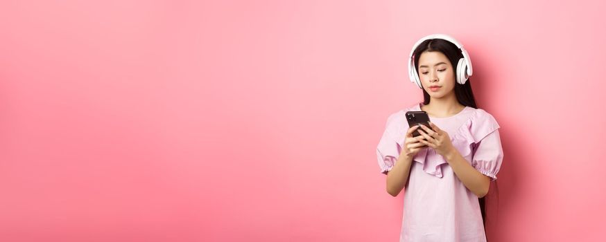 Young girl listening music in headphones and chatting on mobile phone, standing in dress against pink background.