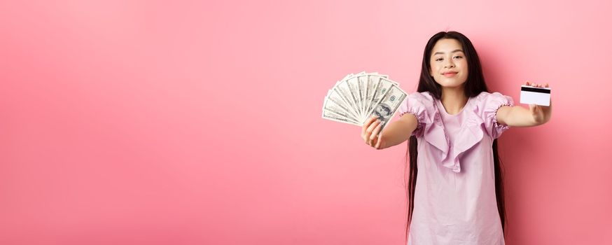 Beautiful asian woman showing plastic credit card and dollar bills, smiling pleased, standing against pink background.