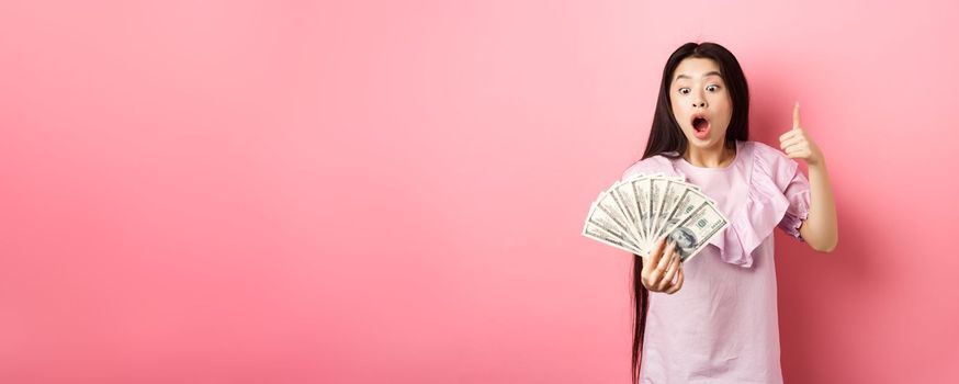 Excited teen girl holding big amount of money, showing dollar bills and thumbs up, standing amazed on pink background.