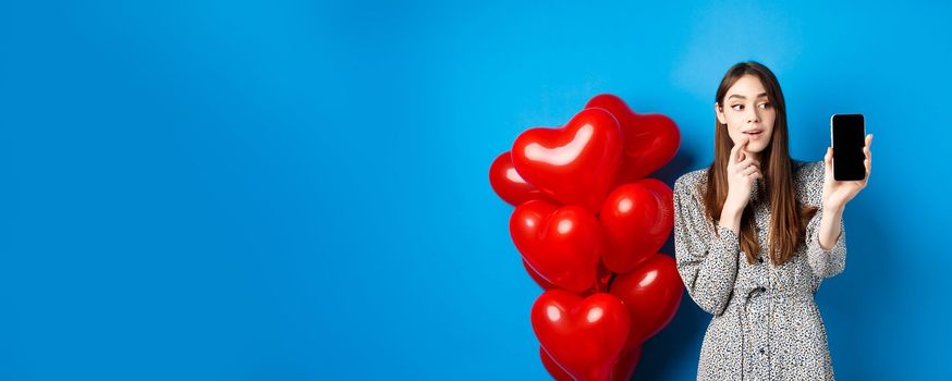 Valentines concept. Beautiful girl looking and showing mobile screen, checking out online shopping offer on lovers day, standing on blue background.