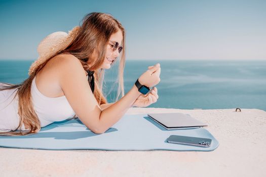 Successful business woman in yellow hat working on laptop by the sea. Pretty lady typing on computer at summer day outdoors. Freelance, travel and holidays concept.