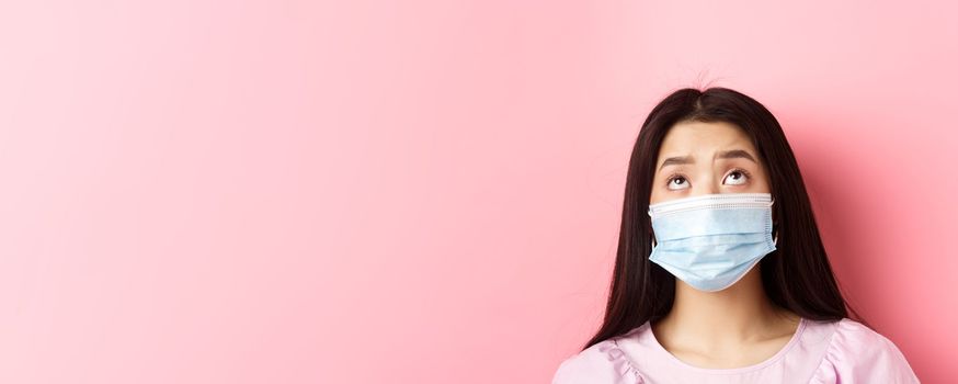 Covid-19, pandemic and quarantine concept. Close-up of sad asian woman in medical mask looking up upset, standing gloomy against pink background.