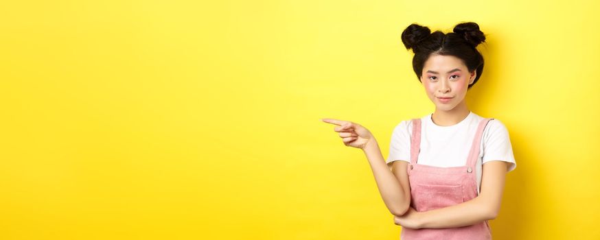 Beautiful teenage asian girl with bright makeup, pointing finger left at banner and smiling, yellow background.