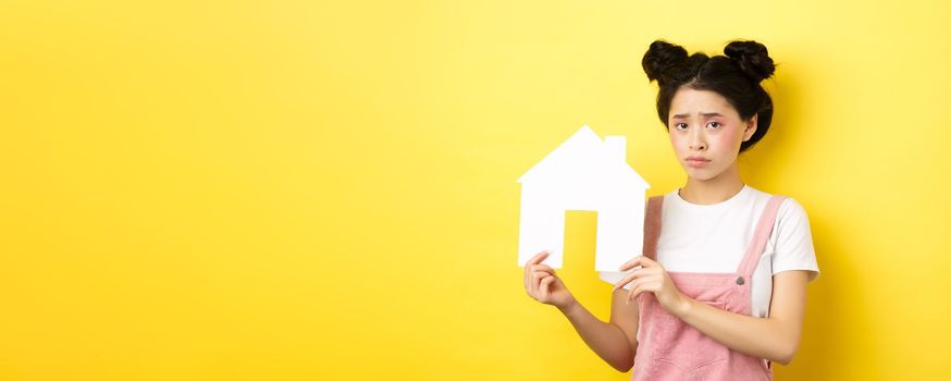 Real estate and family concept. Sad cute asian girl with bright makeup, frowning and feel upset, showing paper house cutout, standing on yellow background.