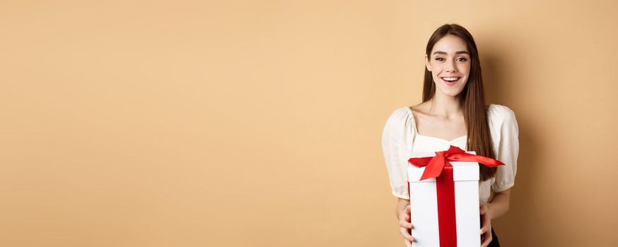 Happy valentines day. Cheerful smiling girl giving surprise gift to lover, looking at camera and holding present in box, standing on beige background.