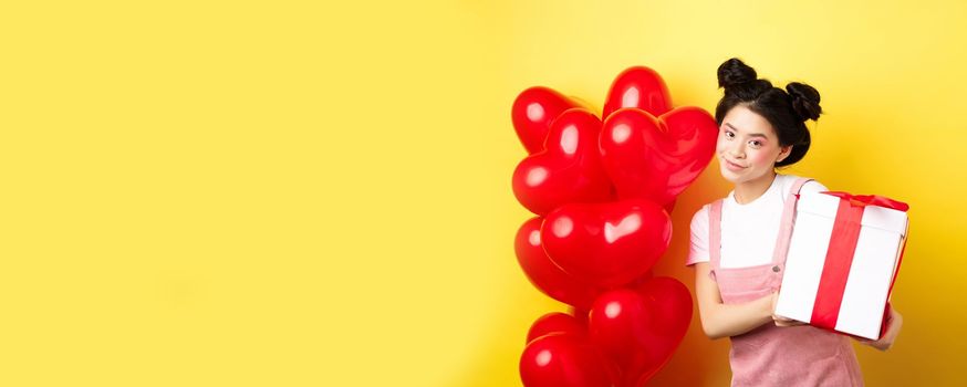 Happy Valentines day. Beautiful and stylish asian woman posing near heart balloons with lovely box with gift from lover, standing over yellow background.