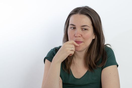 close-up of a young woman, brunette, long-haired, pensive looking at the ground with her finger in her mouth on a white background