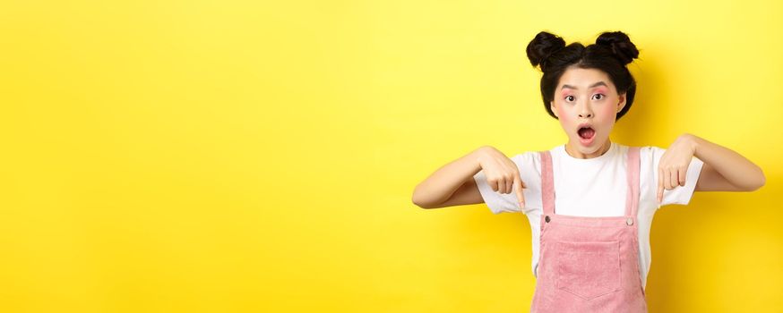 Surprised asian woman with glamour makeup and hairstyle, drop jaw and pointing down amazed, standing against yellow background.