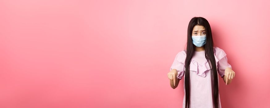 Healthy people and covid-19 pandemic concept. Sad asian woman pointing fingers down, frowning upset, standing against pink background.