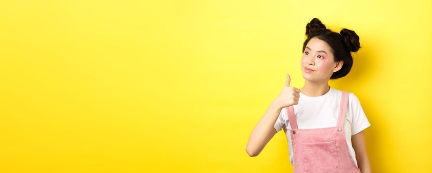 Glamour asian girl with pink makeup and summer clothes, looking left at logo with thumb up, approve and praise, standing on yellow background.