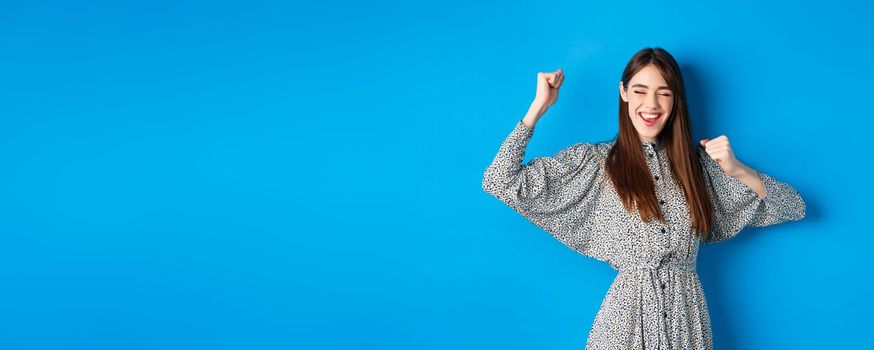 Excited smiling girl showing tongue and dancing with raised hands, celebrating victory or achievement, winning prize, standing happy on blue background.