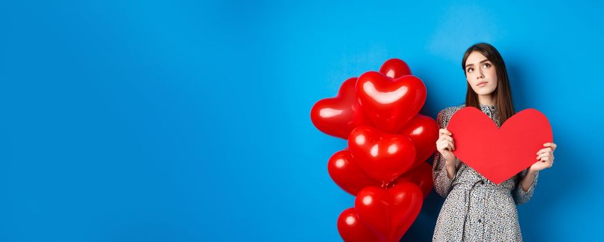 Valentines day. Dreamy pretty lady in dress, holding big red heart cutout and searching for true love, looking up pensive, standing near holiday balloons on blue background.