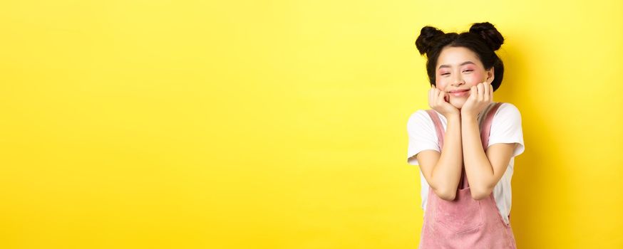 Cute happy asian woman with bright makeup and dungarees, leaning satisfied on hands and smiling, standing on yellow background.