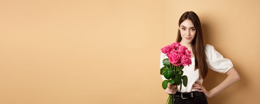 Valentines day. Beautiful girlfriend holding pink roses and looking at camera. Young woman receive flowers from her date, standing on beige background.