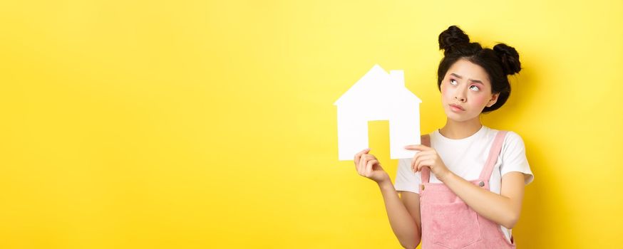 Real estate and family concept. Thoughtful asian girl dream of house, showing paper home cutout and look at upper left corner pensive, yellow background.