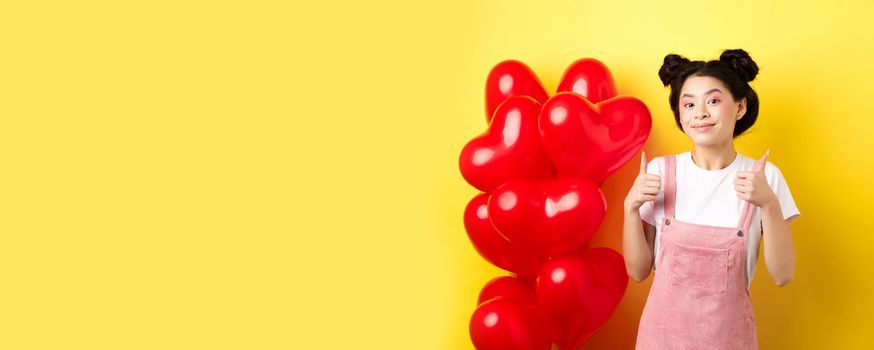 Cute teenage asian girl showing thumbs up, waiting for Valentines day near red heart balloons, wearing outfit for romantic date, yellow background.