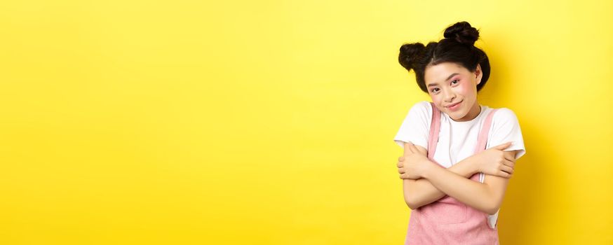 Summer lifestyle concept. Cute asian beauty girl with makeup, hugging herself and smiling romantic, standing tender on yellow background.