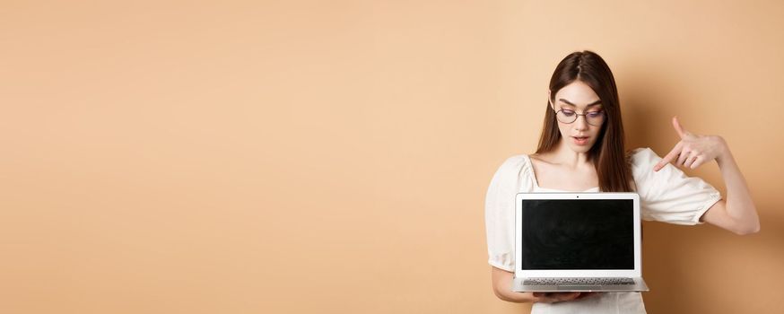 Curious girl in glasses pointing at laptop screen, check out online deal, demonstrate project on computer, standing on beige background.