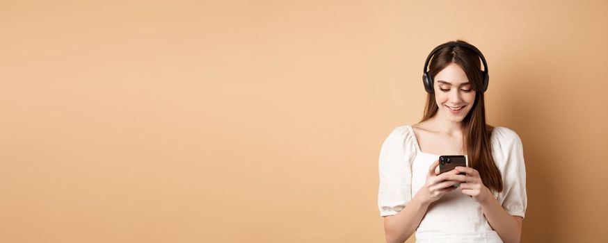 Smiling woman texting message on phone and listening music in wireless headphones, watching video on smartphone, beige background.