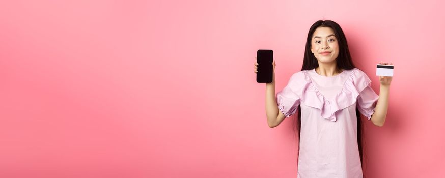 Cheerful asian girl showing empty smartphone screen and credit card, shopping online, standing on pink background.