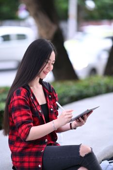Portrait of Asian woman in street style clothes sitting outdoor and using digital tablet.